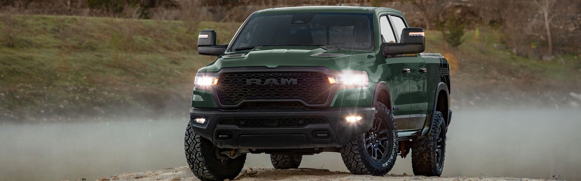 Dark green Ram pickup truck with illuminated headlights and fog lights on a dusty path.